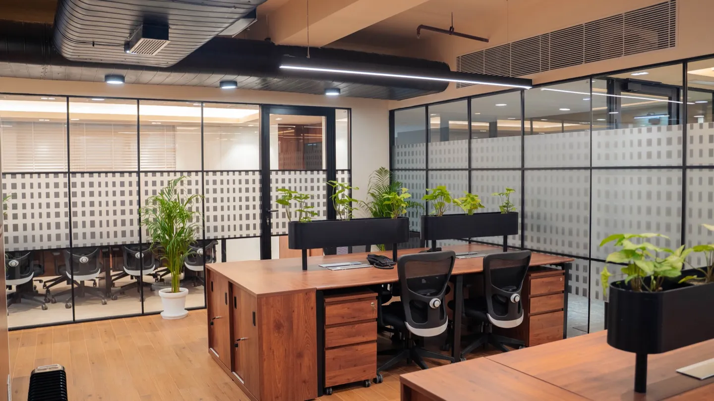 Wooden desk cluster with mesh chairs, planters and glass-walled cabins — AIHP managed office, Gurgaon